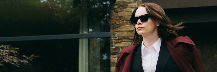 Fotograma de la película Bugonia de Yorgos Lanthimos. Vemos a Michelle, interpretada por Emma Stone. Lleva gafas de sol negras, traje y un abrigo rojo. Tiene el pelo castaño y ondea un poco al viento. Se la ve rica y poderosa. Detrás se ve una casa lujosa y cara. Ella está en el lado derecho de la imagen y mira hacia algún lugar fuera de plano a la izquierda con cara seria y desafiante.