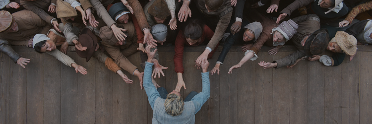 Fotograma de la película Hamnet. Plano cenital. Vemos a un actor de teatro sobre el escenario, de rodillas con las manos extendidas al público. Las personas del público se estiran y alargan las manos hacia él.