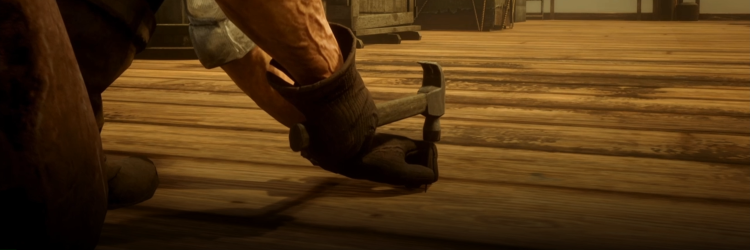 Fotograma del cortometraje Hardly working del colectivo Total Refusal. Vemos a un NPC del videojuego Red Dead Redepmtion. Está de espaldas a la cámara y agachado. En una mano tiene un martillo y con la otra sujeta un clavo contra el suelo. Lleva unos guantes gruesos de trabajo. Va a clavar el clavo en el suelo de tablas de madera sobre el que está. En el fondo se vislumbran unas grandes cajas de madera que parecen transportar mercancías.
