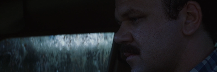 Fotograma de la película Magnolia de Paul Thomas Anderson. Vemos un primer plano de la cara del agente de policía Jim Kurring, interpretado por John C. Reilly, de perfil. Mira ligeramente hacia abajo y tiene una cara taciturna y pensativa. Está dentro de un coche y detrás de él vemos una ventana muy sucia. Él tiene un tupido bigote y una camisa de cuadros azul y blanca.