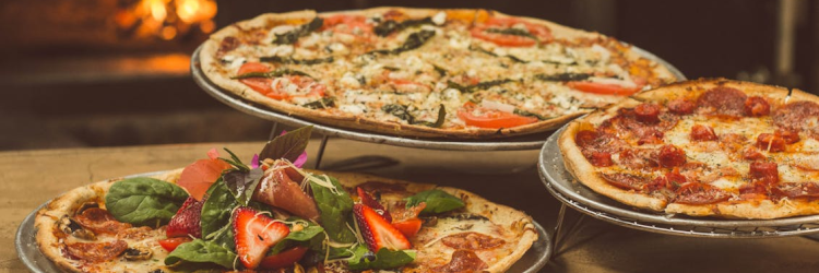 Fotografía de tres pizzas sobre una mesa de madera. La más cercana está sobre un plato metálico, las otras dos sobre soportes metálicos con patas, lo que crea una composición triangular con las pizzas a tres alturas distintas. En el fondo se vislumbra la luz de un horno de leña, pero está muy desenfocada.