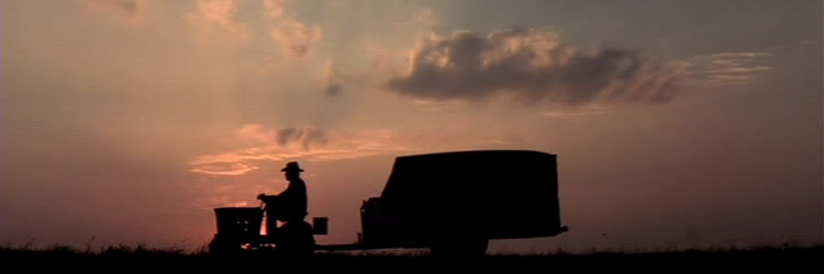 Fotograma de la película The Straight story de David Lynch. Se ve la silueta de perfil de un hombre mayor con sombrero vaquero que conduce un cortacesped. El cortacesped arrastra un remolque más grande que él. Al fondo se ve el cielo del atardecer, con un color más rojizo cuanto más cerca de la línea del horizonte, con unas pocas nubrs sueltas.